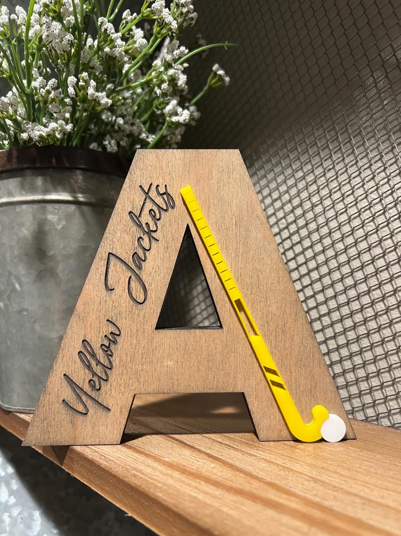 May include: Wooden letter A with the words "Yellow Jackets" engraved on it. A yellow field hockey stick is placed on the letter A.
