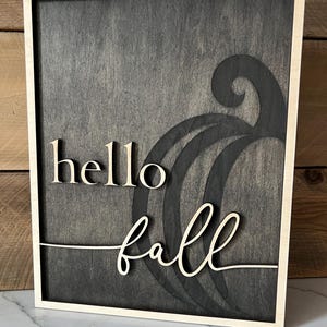 Może przedstawiać: Drewniany znak z czarnym tłem i jasną drewnianą ramą. Znak głosi "hello fall" ozdobnym fontem. Słowo "fall" jest nałożone na stylizowany kształt dyni.