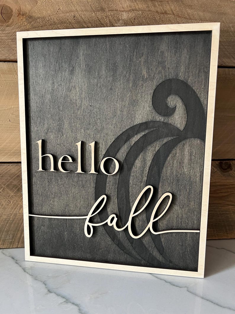 Może przedstawiać: Drewniany znak z czarnym tłem i jasną drewnianą ramą. Znak głosi "hello fall" ozdobnym fontem. Słowo "fall" jest nałożone na stylizowany kształt dyni.