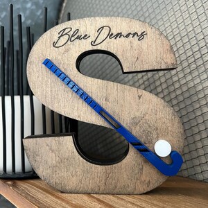 May include: Wooden letter S with a blue hockey stick and white puck, with the words "Blue Demons" engraved on the top.