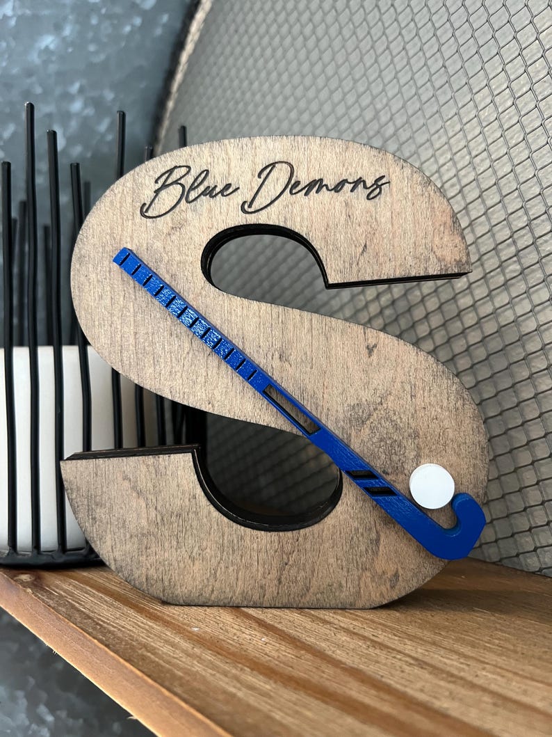 May include: Wooden letter S with a blue hockey stick and white puck, with the words "Blue Demons" engraved on the top.
