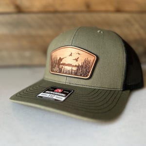 May include: A green and black trucker hat with a brown leather patch depicting a hunter and ducks in a marsh. The patch has the text "Richardson 112" on it.