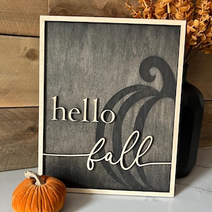 Może przedstawiać: Drewniany znak z czarnym tłem i białą drewnianą ramą. Znak głosi "hello fall" kursywą. Stylizowana dynia jest wyrzeźbiona w drewnie za tekstem.