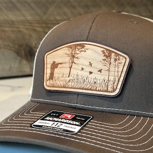 May include: A brown trucker hat with a white mesh back and a brown brim. The hat features a wooden patch with a hunting scene, including a hunter, dog, trees, and flying birds. The patch is rectangular with rounded corners.