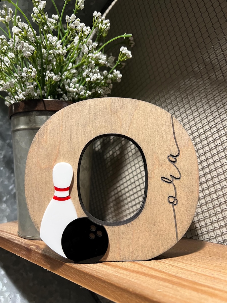 Pu&ograve; includere: Lettera O in legno con un biril da bowling bianco e rosso e una palla da bowling nera con tre fori. La lettera &egrave; dipinta di marrone e ha la scritta "oh la".