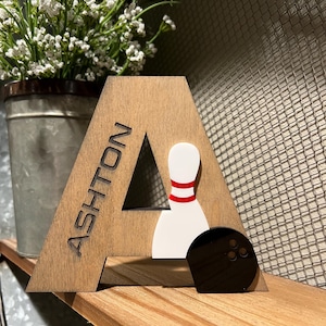 Pu&ograve; includere: Lettera A in legno con il nome Ashton inciso su di essa. La lettera &egrave; decorata con un birilli da bowling bianco con strisce rosse e una palla da bowling nera.