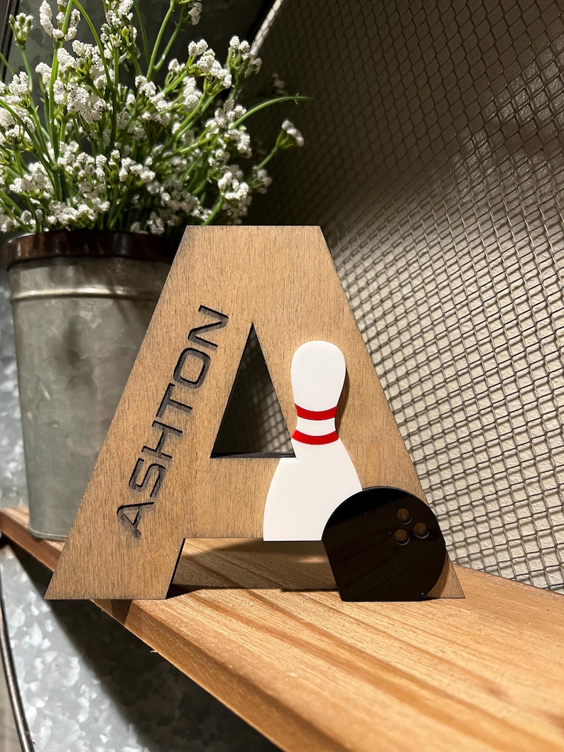 Pu&ograve; includere: Lettera A in legno con il nome Ashton inciso su di essa. La lettera &egrave; decorata con un birilli da bowling bianco con strisce rosse e una palla da bowling nera.