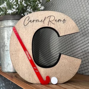 May include: Wooden letter C with a red hockey stick and a white puck. The letter is painted brown and has the text "Carmel Rams" engraved on it.