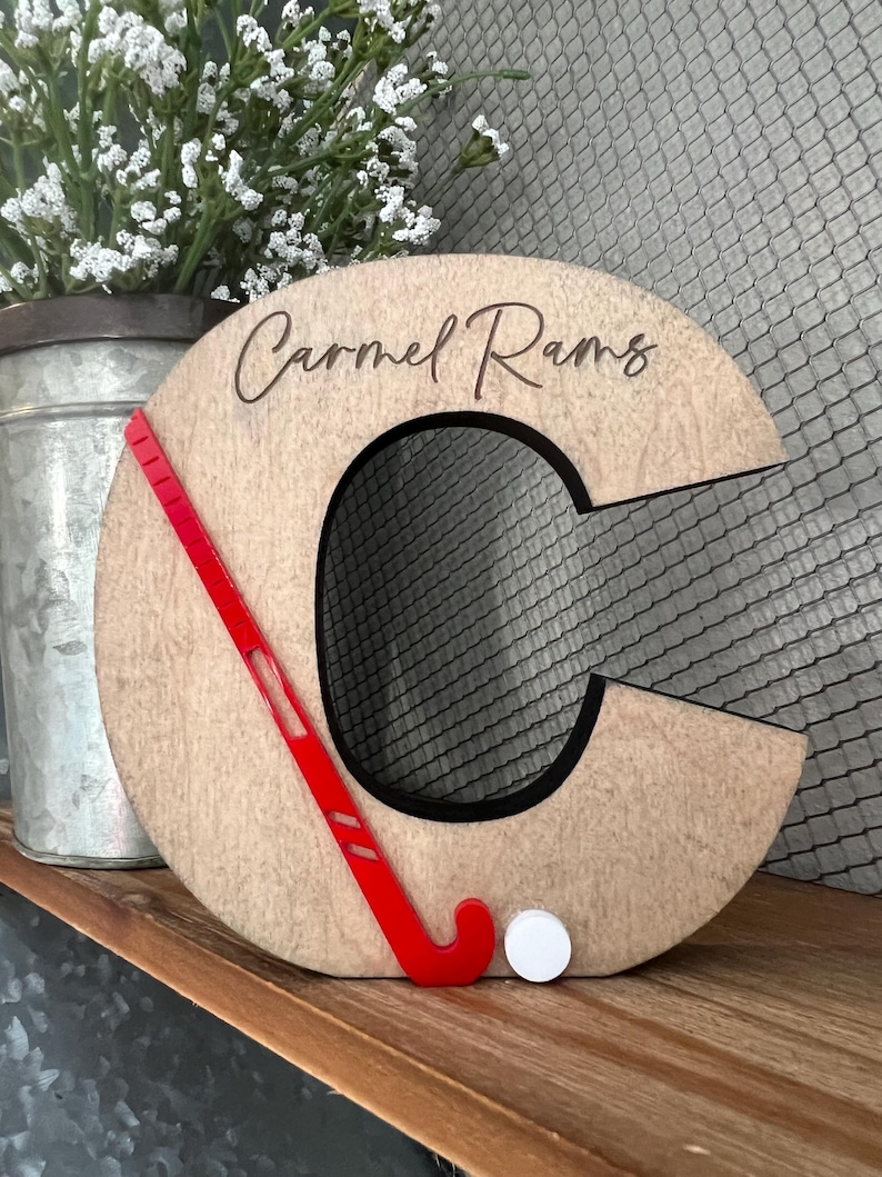 May include: Wooden letter C with a red hockey stick and a white puck. The letter is painted brown and has the text "Carmel Rams" engraved on it.