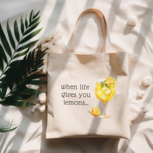 May include: A white canvas tote bag with the text "when life gives you lemons..." and a watercolor illustration of a glass of lemonade with ice cubes and a lemon slice.