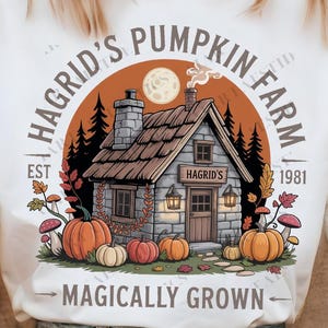 Puede incluir: Camiseta blanca con un diseño de una cabaña de piedra, calabazas y el texto "Hagrid's Pumpkin Farm" y "Magically Grown". El diseño incluye una luna llena y árboles.