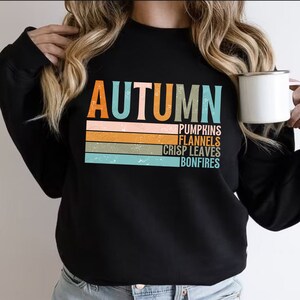 May include: Black sweatshirt with a vintage-style graphic that says "AUTUMN" with the words "pumpkins, flannels, crisp leaves, bonfires" stacked underneath.