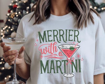 May include: White t-shirt with a green and red graphic that says "Merrier with a Martini" and a martini glass with a red drink and a green olive.