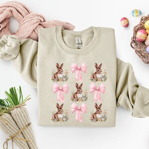 May include: A beige sweatshirt with a repeating pattern of brown bunnies and pink bows. The bunnies are surrounded by small flowers.