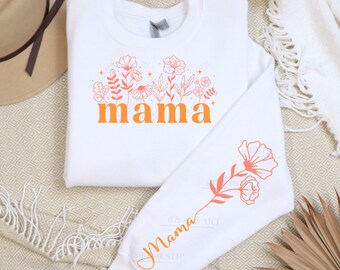 May include: White sweatshirt with a floral design and the word "mama" in orange lettering. The sweatshirt has a long sleeve with a single flower design on the cuff.