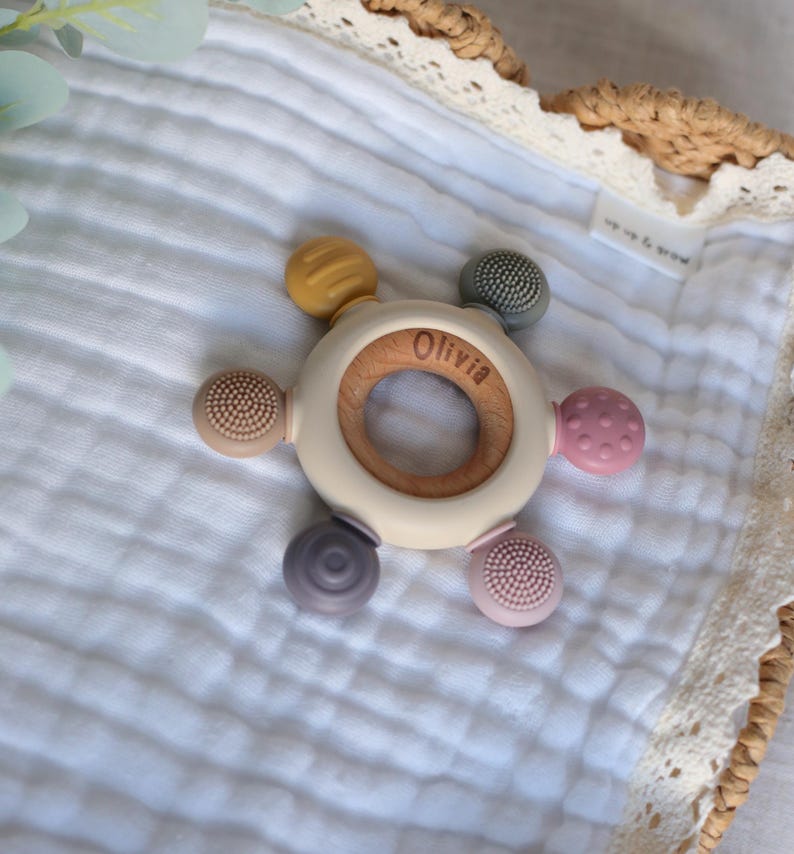 Pu&ograve; includere: Un anello da dentizione in legno con un anello esterno in silicone bianco e cinque diversi attacchi in silicone testurizzati in tonalit&agrave; di viola, rosa, marrone e giallo. L'anello &egrave; inciso con il nome "Olivia".