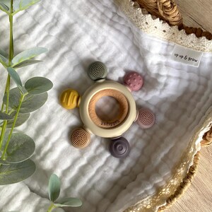 Pu&ograve; includere: Un mordedor per neonati beige e marrone con un anello di legno al centro e cinque attacchi in silicone testurizzati in vari colori, tra cui verde, giallo, rosa e viola. Il mordedor &egrave; su una coperta di mussola bianca con il testo "up up & grow" sulla coperta.