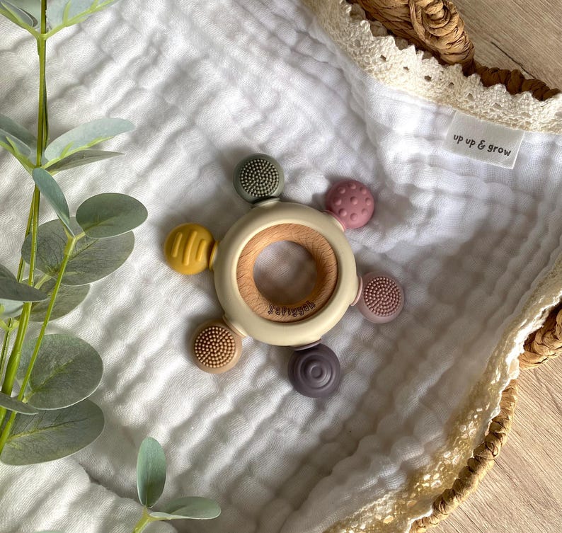 Pu&ograve; includere: Un mordedor per neonati beige e marrone con un anello di legno al centro e cinque attacchi in silicone testurizzati in vari colori, tra cui verde, giallo, rosa e viola. Il mordedor &egrave; su una coperta di mussola bianca con il testo "up up & grow" sulla coperta.