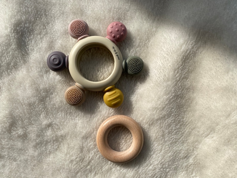 Pu&ograve; includere: Un anello da dentizione bianco con cinque diversi attacchi in silicone testurizzati in rosa, viola, giallo e verde. L'anello &egrave; su una superficie bianca e pelosa. C'&egrave; un anello di legno separato sulla superficie.