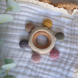 Pu&ograve; includere: Un anello da dentizione in legno con un anello esterno in silicone bianco e cinque diversi attacchi in silicone testurizzati. L'anello &egrave; inciso con il nome "Olivia".