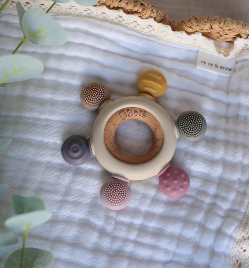 Pu&ograve; includere: Un anello da dentizione in legno con un anello esterno in silicone bianco e cinque diversi attacchi in silicone testurizzati. L'anello &egrave; inciso con il nome "Olivia".