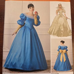 Könnte beinhalten: Simplicity-Schnittmuster 1728 D5 zeigt ein Prinzessinnenkostüm. Das Muster beinhaltet ein blaues Kleid mit Puffärmeln, eine gelbe Schleife und eine Krone. Das Bild zeigt auch ein Model, das das Kostüm trägt, und eine Zeichnung eines ähnlichen Kleides.