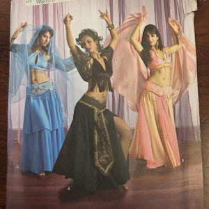 Puede incluir: Patrón de estudio de danza Simplicity con tres bailarinas de danza del vientre con trajes. Los trajes son en tonos azules, negros y rosas, con detalles intrincados. El patrón incluye el texto "Simplicity the dance studio" y "Andrea Schewe".