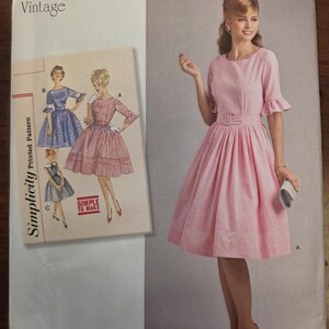 Puede incluir: Patrón de costura Simplicity vintage de los años 1960. El patrón presenta un vestido rosa con cintura ceñida y falda acampanada. El sobre del patrón incluye ilustraciones del vestido y el texto "SIMPLE TO MAKE".