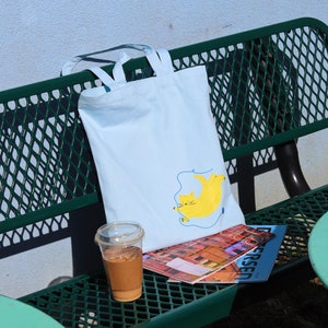 May include: A light blue tote bag with a yellow cat illustration and a blue string. The bag is sitting on a green metal bench with a magazine and a cup of iced coffee.