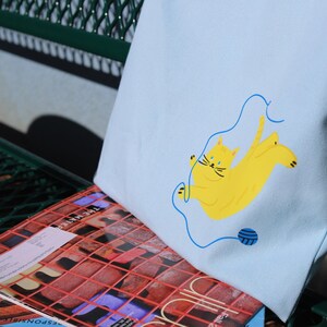 May include: A light blue tote bag featuring an illustration of a yellow cat. The cat is tangled in blue yarn and holding a ball of yarn.