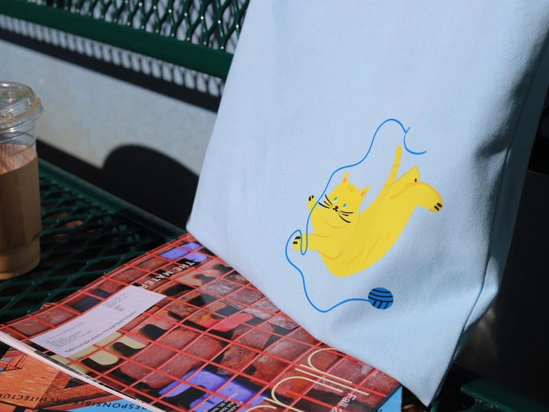 May include: A light blue tote bag featuring an illustration of a yellow cat. The cat is tangled in blue yarn and holding a ball of yarn.