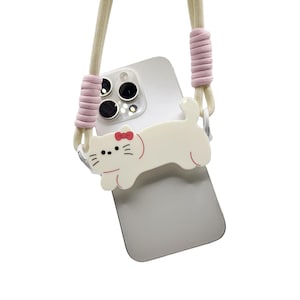 May include: A white phone case with a pink and white cat design. The case has a pink and white strap attached to it.