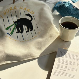 以下が含まれることがあります： 黒い猫のイラストと「私は一生懸命働くので、人間はより良い生活を送ることができる」というテキストが書かれた、ベージュのキャンバス地のトートバッグです。