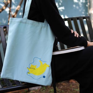 May include: A light blue tote bag featuring a yellow cat design. The cat is playing with a ball of yarn.