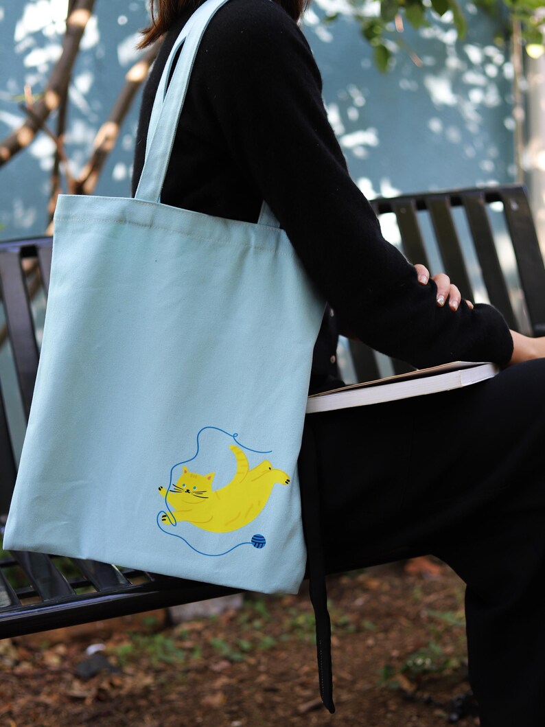May include: A light blue tote bag featuring a yellow cat design. The cat is playing with a ball of yarn.