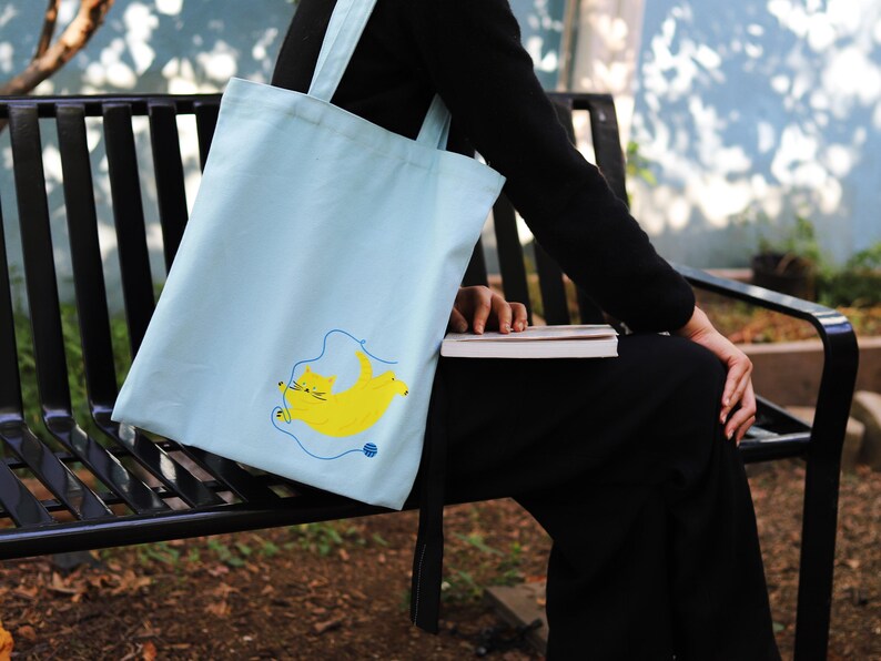 May include: A light blue tote bag featuring a yellow cat design. The cat is playing with a ball of yarn. The bag is resting on a black metal bench.