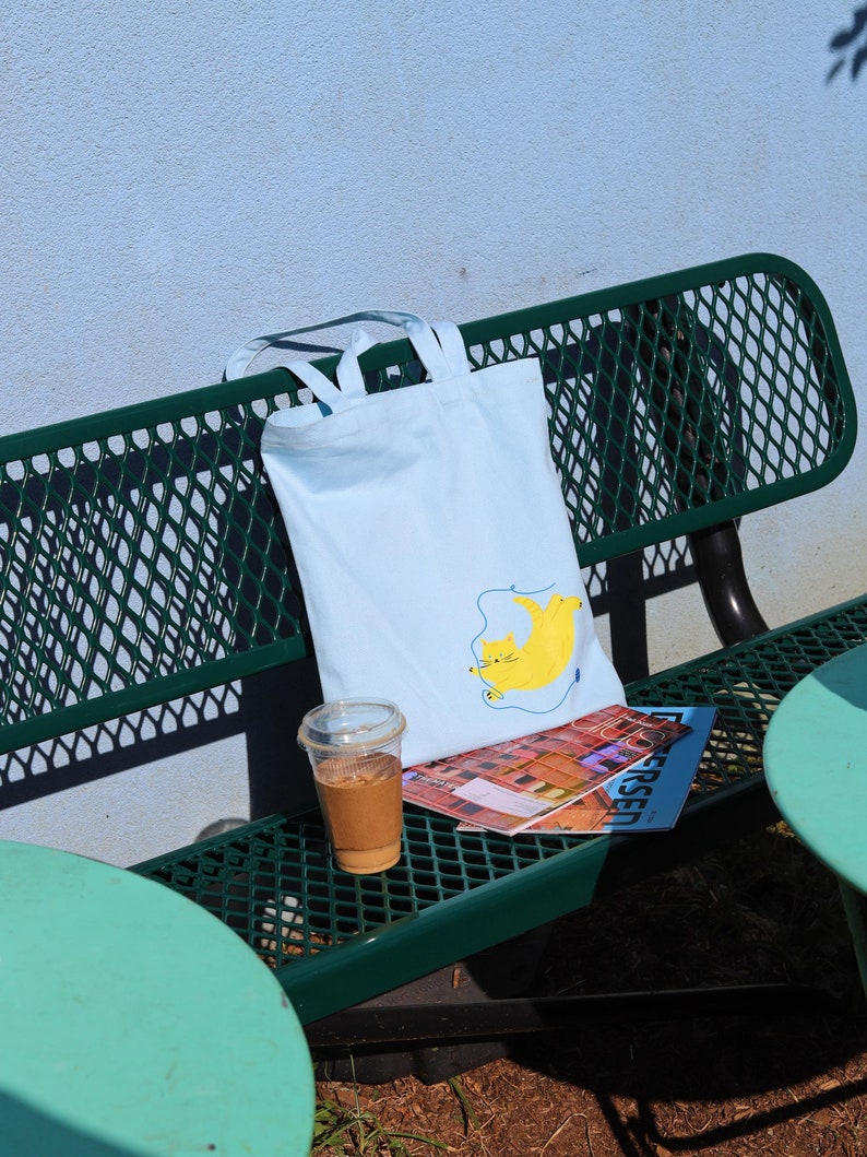 May include: A light blue tote bag with a yellow cat illustration and a blue string. The bag is sitting on a green metal bench with a magazine and a cup of iced coffee.