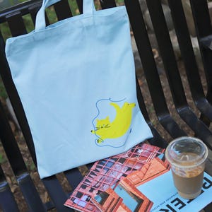 May include: A light blue tote bag featuring a yellow cat illustration. The cat is depicted with a blue string attached to its tail. The bag is resting on a black metal bench alongside a magazine and a glass of iced coffee.