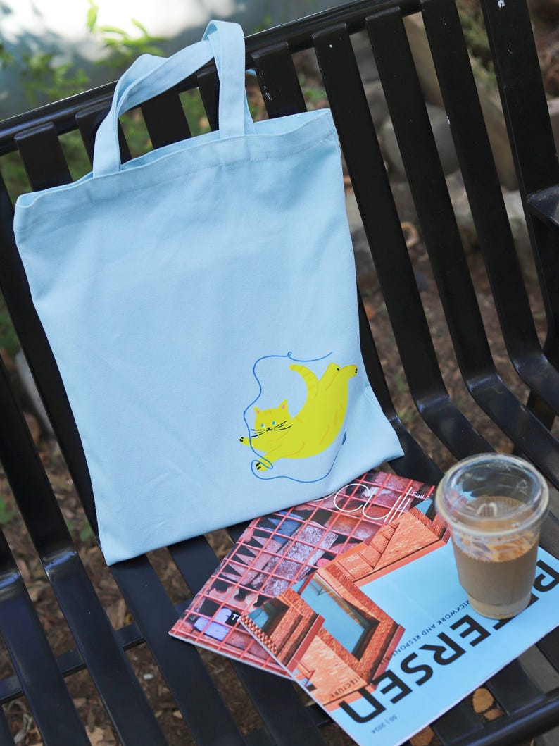 May include: A light blue tote bag featuring a yellow cat illustration. The cat is depicted with a blue string attached to its tail. The bag is resting on a black metal bench alongside a magazine and a glass of iced coffee.
