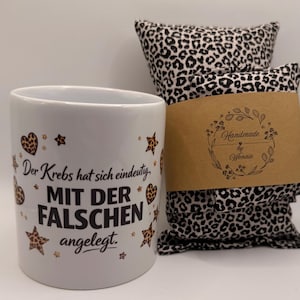 May include: A white ceramic mug with the text "Der Krebs hat sich eindeutig MIT DER FALSCHEN angelegt" and leopard print hearts and stars. Next to it are two leopard print pillows with a brown paper label that says "Handmade by Wonnle".