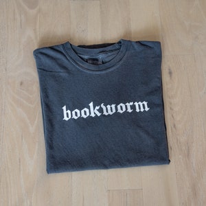 Op de afbeelding: Een donkergrijs T-shirt met het woord "bookworm" in wit gedrukt in een gotisch lettertype.