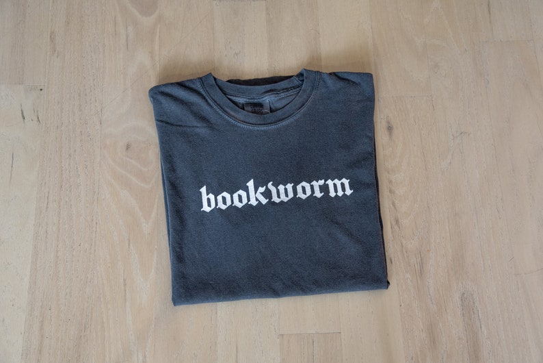 Op de afbeelding: Een donkergrijs T-shirt met het woord "bookworm" in wit gedrukt in een gotisch lettertype.