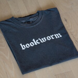 Op de afbeelding: Een donkergrijs T-shirt met het woord "bookworm" in wit gedrukt in een gotisch lettertype.
