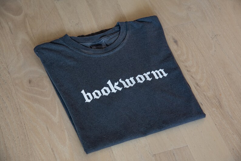 Op de afbeelding: Een donkergrijs T-shirt met het woord "bookworm" in wit gedrukt in een gotisch lettertype.