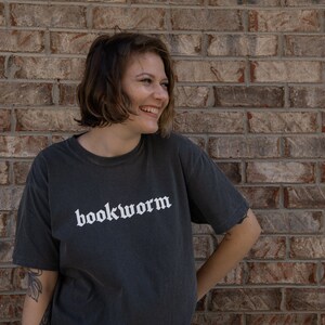 Op de afbeelding: Een persoon draagt een donkergrijs T-shirt met het woord "bookworm" in wit op de voorkant. De persoon staat voor een bakstenen muur.