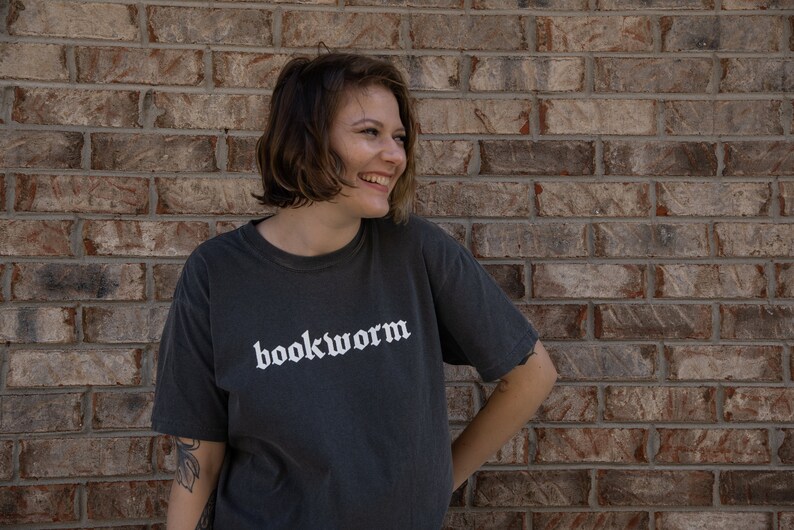Op de afbeelding: Een persoon draagt een donkergrijs T-shirt met het woord "bookworm" in wit op de voorkant. De persoon staat voor een bakstenen muur.