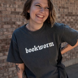 Op de afbeelding: Een vrouw draagt een donkergrijs T-shirt met het woord "bookworm" in wit op de voorkant. Ze glimlacht en kijkt naar de zijkant. Ze heeft kort bruin haar en draagt een zwart shirt onder het T-shirt.
