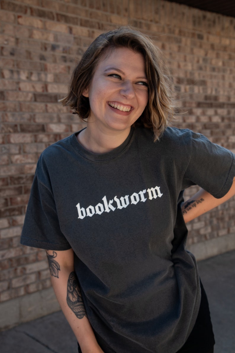 Op de afbeelding: Een vrouw draagt een donkergrijs T-shirt met het woord "bookworm" in wit op de voorkant. Ze glimlacht en kijkt naar de zijkant. Ze heeft kort bruin haar en draagt een zwart shirt onder het T-shirt.