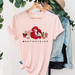 Op de afbeelding: Roze T-shirt met een afbeelding van Ariel uit De Kleine Zeemeermin, Mickey Mouse en andere Disney-figuren. De tekst "BEST DAY EVER" is onder de afbeelding gedrukt.