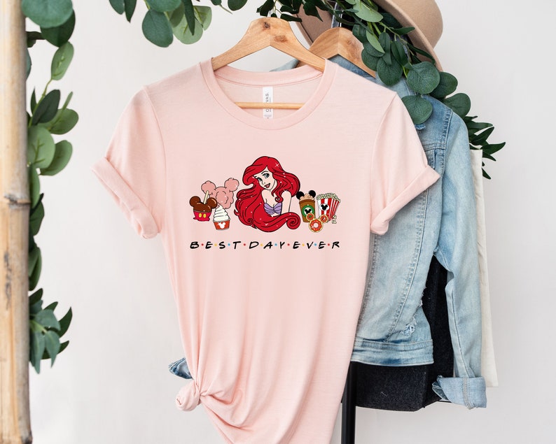 Op de afbeelding: Roze T-shirt met een afbeelding van Ariel uit De Kleine Zeemeermin, Mickey Mouse en andere Disney-figuren. De tekst "BEST DAY EVER" is onder de afbeelding gedrukt.